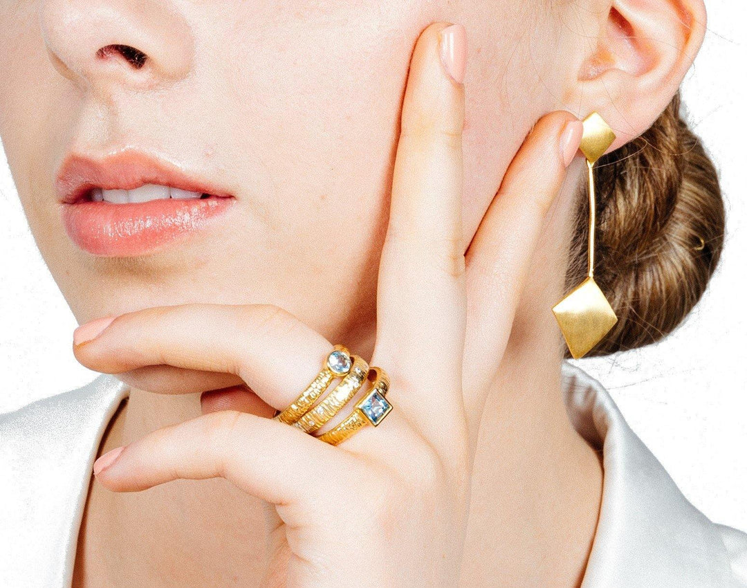 Close-up of a woman’s face and hand. She is wearing gold jewelry: two gold rings with gemstones on her fingers and an Art Deco Earrings In Gold Vermeil by Gallardo & Blaine Designs featuring a geometric design in gold vermeil. She is touching her face with her fingers, and her hair is styled in a bun. The background is white.