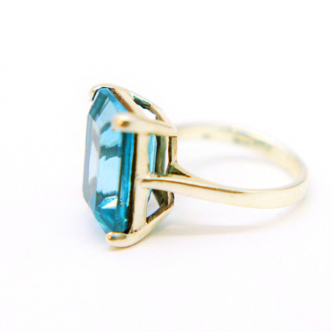 A close-up image of the Stellar Ring in Hydro Blue Topaz by Gallardo & Blaine Designs, featuring a large, rectangular blue topaz set in a pronged setting. The gemstone is translucent and has a faceted cut, catching the light on its surface. The band is simple and smooth. The background is white.