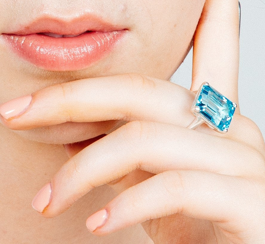 Close-up of a woman's lips and fingers. She is wearing a large, rectangular-cut Stellar Ring in Hydro Blue Topaz by Gallardo & Blaine Designs on her index finger crafted from sterling silver. The nails are manicured with a pale pink color. The background is neutral.