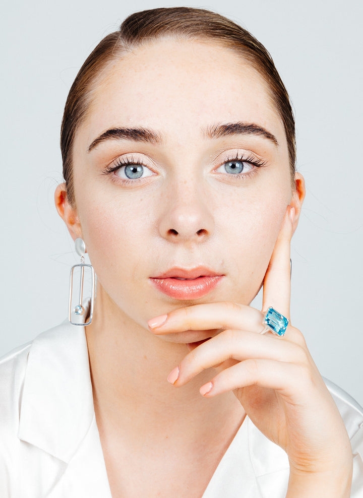 Close-up of a woman with light skin and blue eyes, staring directly at the camera. She has her hair neatly pulled back and rests her chin on her hand, adorned with a Gallardo & Blaine Designs Stellar Ring in Hydro Blue Topaz. She is wearing a white top and pearl earring with a geometric design.