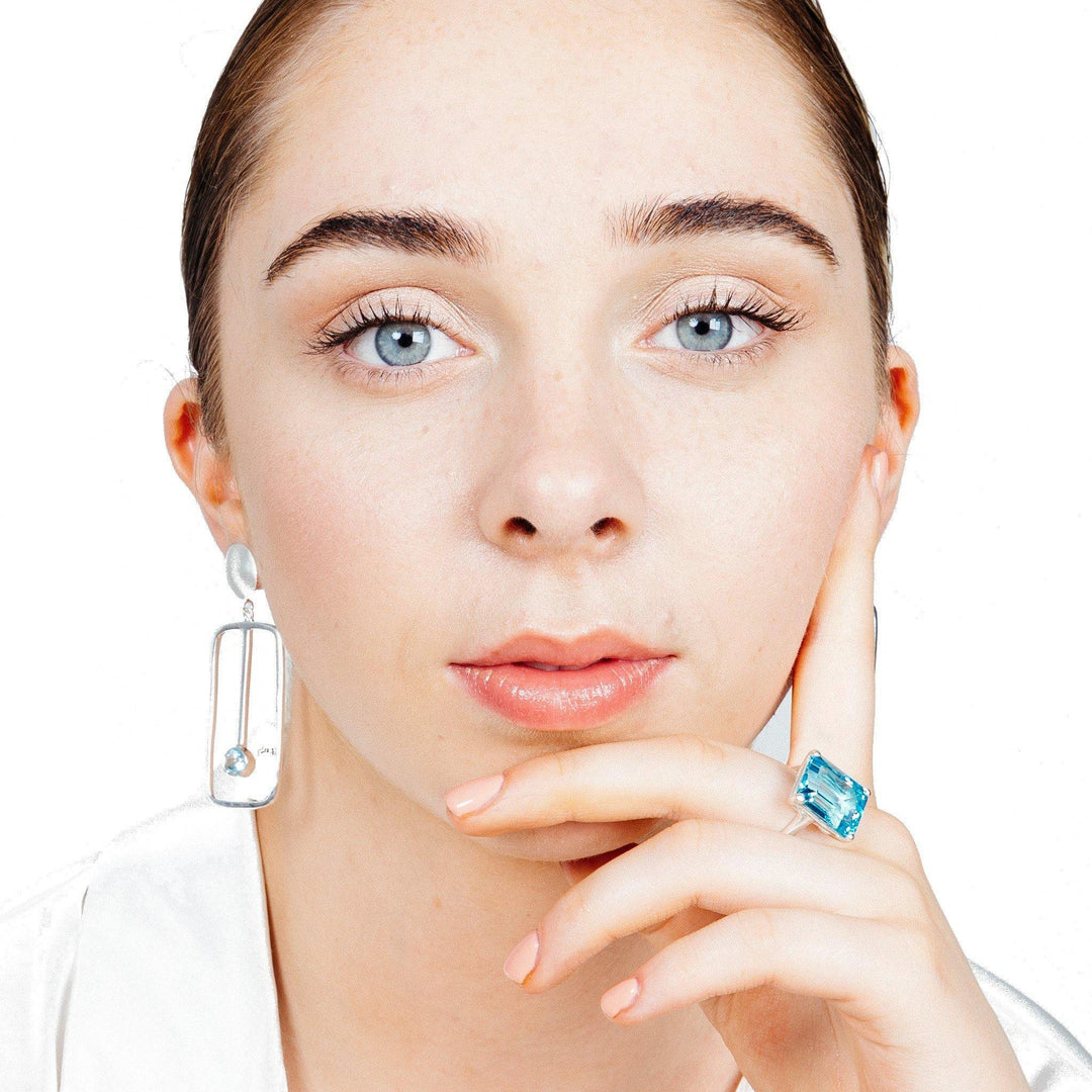 A person poses against a white background. They have light colored eyes and hair that is pulled back, wearing silver Lorelei Earrings Large in various gemstones from Gallardo & Blaine Designs with a small gemstone and a large, rectangular blue ring on their finger. Their hand is resting near their face with a neutral expression.