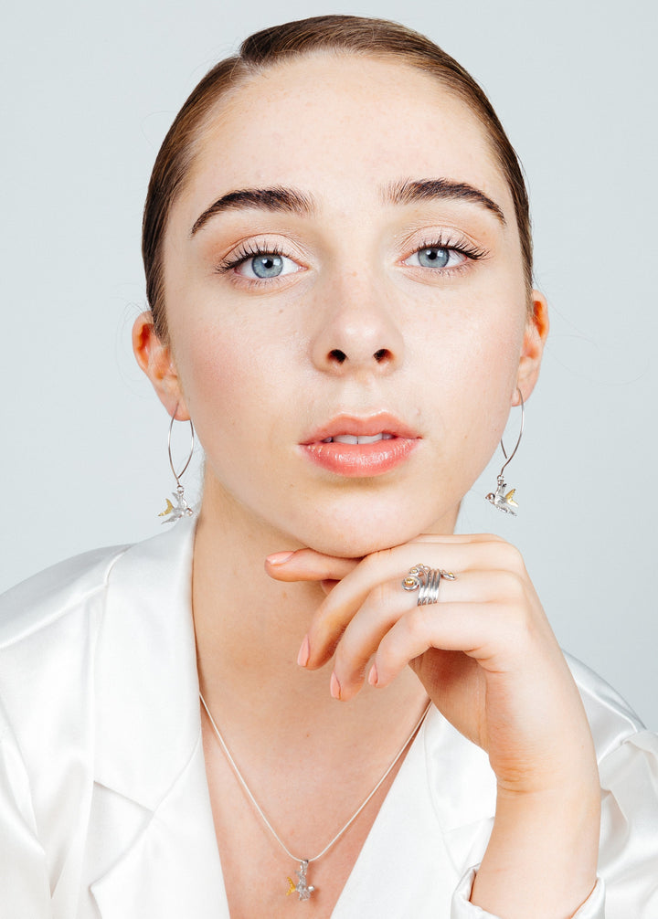 A person with light skin, light blue eyes, and brown hair, pulled back neatly, is wearing silver hoop earrings with small dangling charms, a sterling silver snake chain necklace with a Wildlife Pendant - Swallow With Chain by Gallardo & Blaine Designs, and a silver ring. They rest their chin on their hand and are dressed in a white satin top.