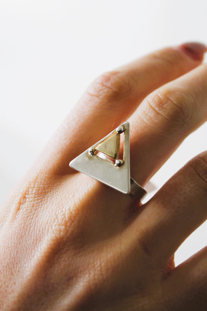 A hand displays the Gallardo & Blaine Designs Triune Ring, a sterling silver piece with a hollow triangle and metallic spheres at each corner, against a soft, light background.