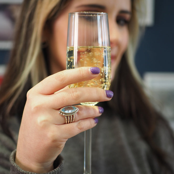 A person with long hair is holding a champagne glass filled with a bubbly drink. They have purple nail polish and are wearing the Tulip Ring in various gemstones by Gallardo & Blaine Designs on their finger. The background is blurred, focusing attention on the glass and hand.