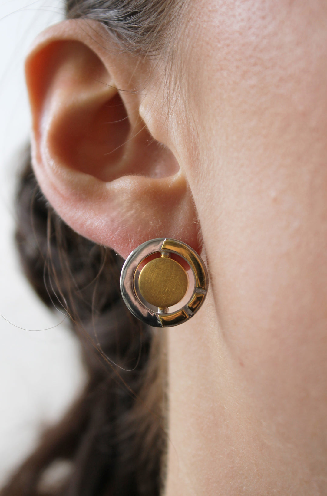 Close-up of a person's ear wearing Urú Studs by Gallardo & Blaine Designs—round stud earrings crafted from gold and sterling silver with a circular design. The background is blurred.