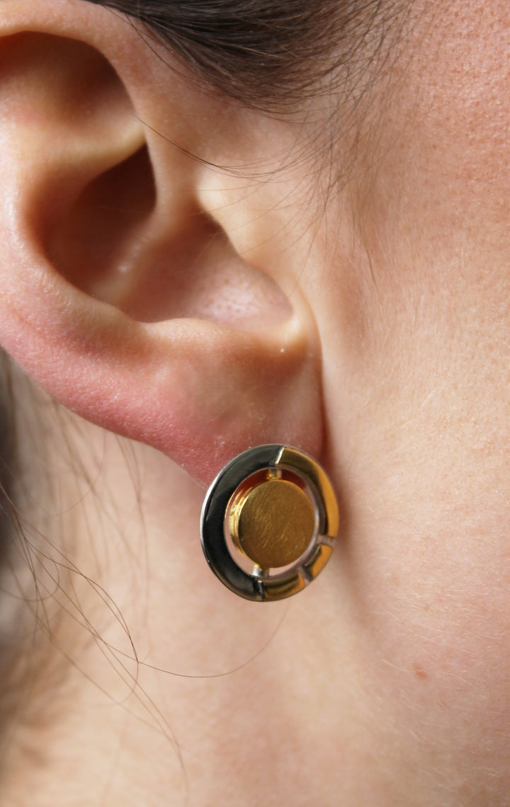 Close-up of a person's ear wearing the Urú Studs by Gallardo & Blaine Designs—round, gold and sterling silver geometric earrings inspired by Celtic motifs with a central gold circle encircled by a sterling silver design.