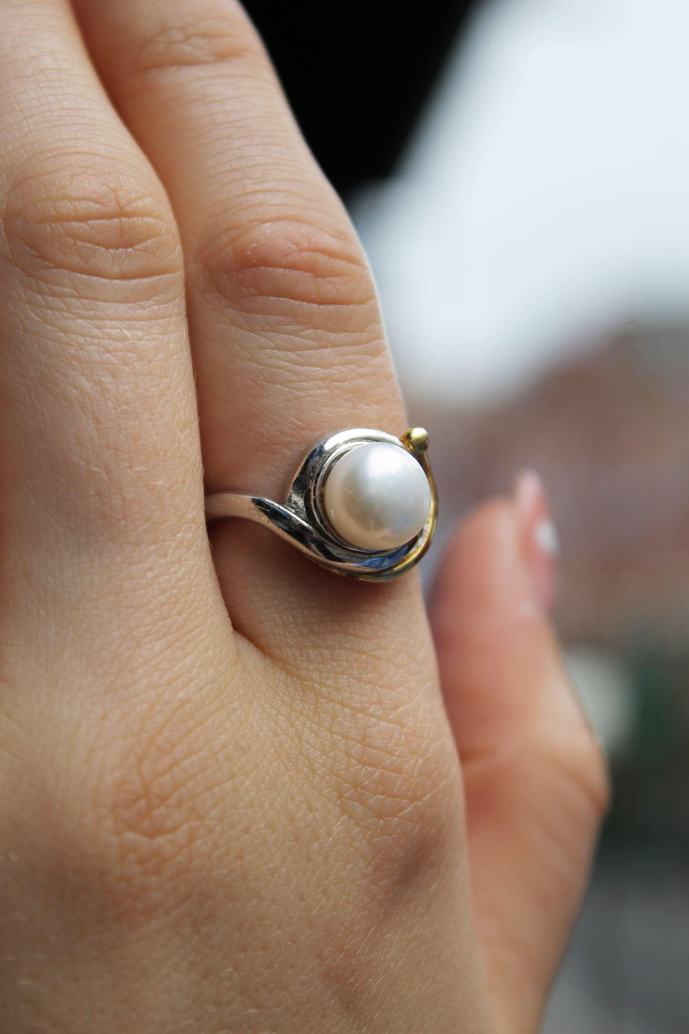 A close-up of a hand wearing the Wave Ring by Gallardo & Blaine Designs, featuring a round white pearl set in silver, against a blurred outdoor background.