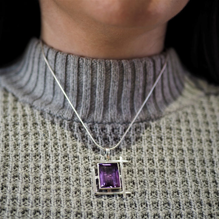 A person is wearing the Gallardo & Blaine Designs Art Deco Pendant in various gemstones featuring a large rectangular purple gemstone. The pendant is set in a simple geometric frame. The individual is dressed in a gray, ribbed turtleneck sweater.