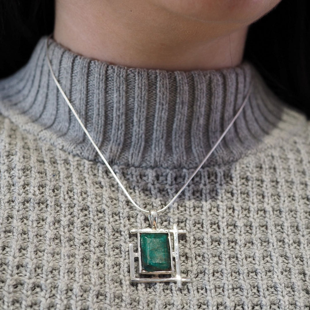 A person wearing a gray, ribbed, high-neck sweater with a sterling silver necklace featuring an Art Deco Pendant in various gemstones by Gallardo & Blaine Designs. The rectangular green stone is set in a geometric silver frame, hanging just below the collar. Only the lower part of their face and neck are visible.