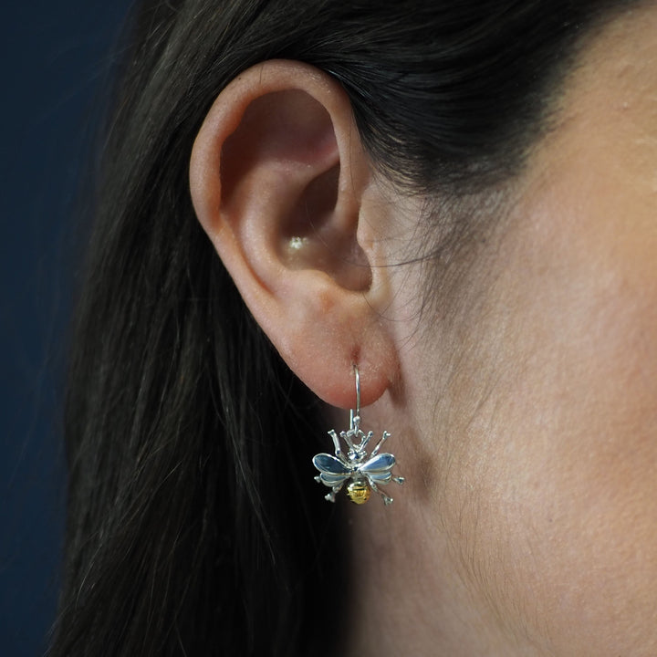 A close-up of a person's ear adorned with Wildlife Bee Dangle Earrings by Gallardo & Blaine Designs. The metallic body, light blue-tinted wings, and small yellow element for the belly create a stunning piece of wildlife-themed jewelry. The solid dark blue background and the partially visible dark hair add to the elegance.