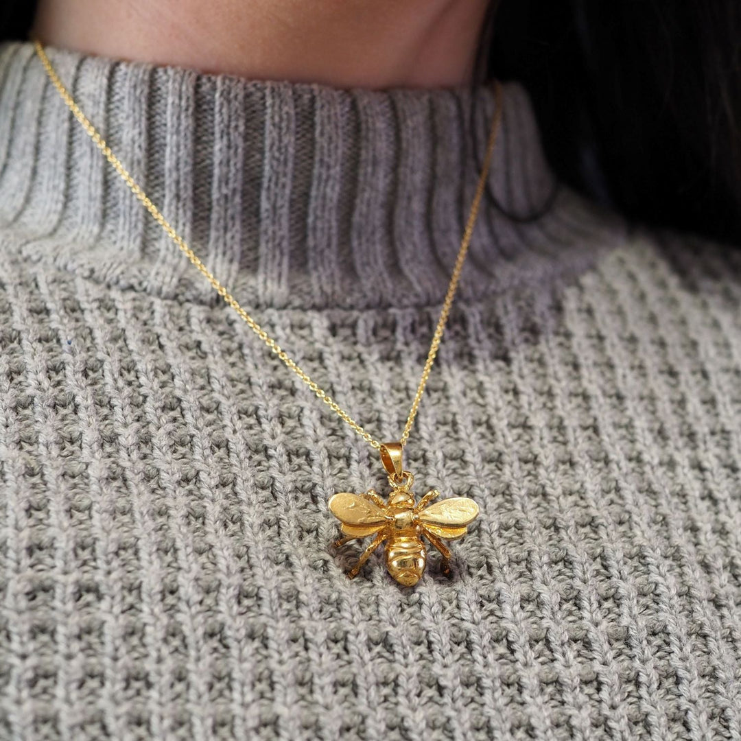 A close-up of a person wearing a gray knit sweater and a gold-plated necklace featuring the **Wildlife Pendant - Large Bee With Chain** by **Gallardo & Blaine Designs**. The bee pendant has intricately carved wings and body, adding texture and sophistication to the accessory.