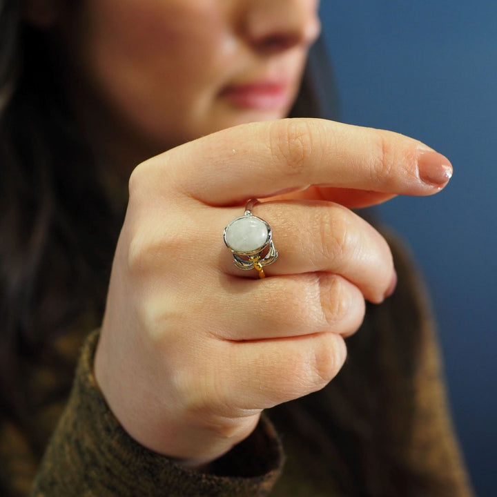 A person holds their hand up, displaying a delicate and dainty Daydream Ring in various gemstones with a round, opaque white stone set in a silver band by Gallardo & Blaine Designs. The person's face is partially visible, focusing on the timeless piece. The background is blue, and the person wears a brown sweater.