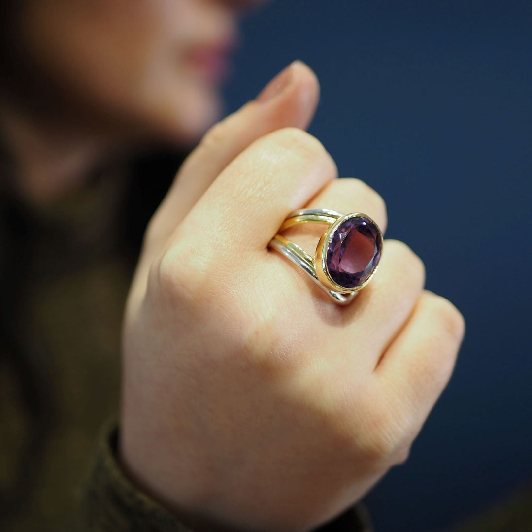 A close-up of a person's hand wearing the Dewberry Ring in various gemstones by Gallardo & Blaine Designs, featuring a chunky statement ring with a large, oval-shaped purple gemstone set in sterling silver. The background is blurred, and the focus is on the striking hand and elegant ring.