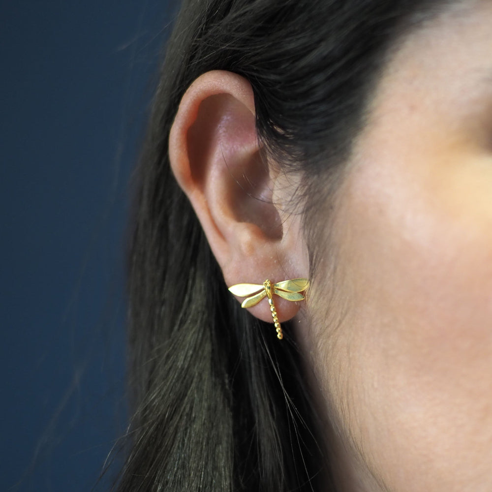 Close-up of a woman's ear adorned with Wildlife Large Dragonfly Studs by Gallardo & Blaine Designs, gold plated for an elegant touch. The background is a soft, solid blue, with only part of her face and hair visible. The earring's intricate details resemble a delicate dragonfly in flight.