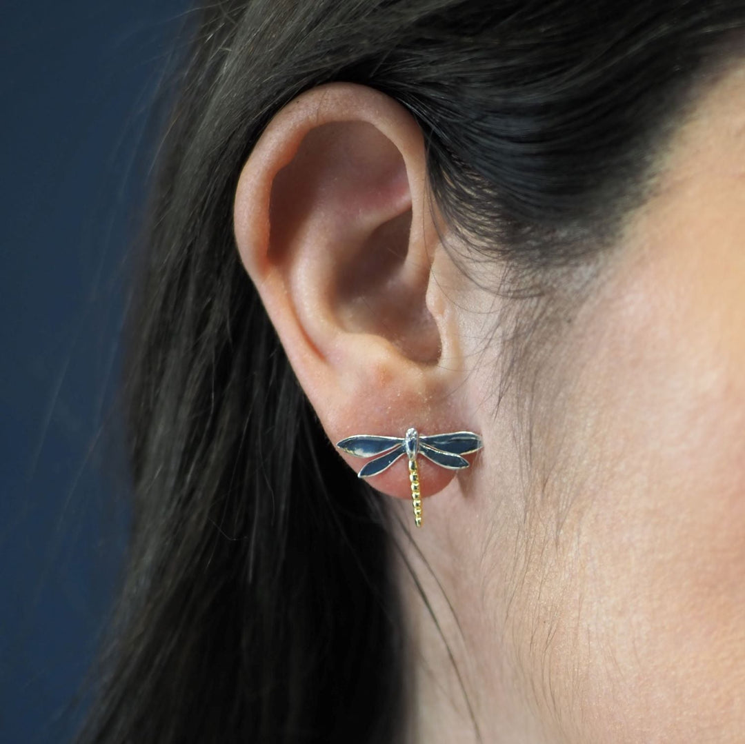 Close-up of a person’s ear wearing Gallardo & Blaine Designs Wildlife Large Dragonfly Studs. The earrings feature a sleek design with blue wings and a gold-colored body. The background is blurred, highlighting the ear and earring.