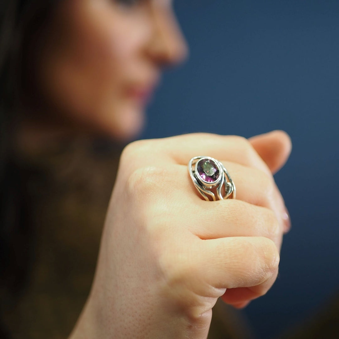 In the foreground, a hand is wearing a Fossil Ring in silver & various gemstones by Gallardo & Blaine Designs. The background is blurred, featuring a person with dark hair and a blue backdrop. The focus is on the adjustable ring.