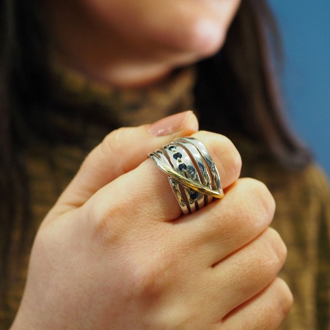 Close-up of a person's hand wearing a chunky statement ring in sterling silver with an intricate design featuring multiple overlapping bands. The person is wearing a brownish sweater, and the background is blurred, emphasizing the Hidden Gems Ring in Silver & Gold by Gallardo & Blaine Designs.