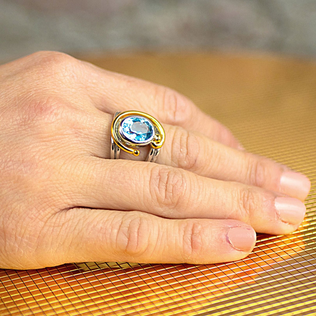 A person's hand with neatly trimmed nails rests on a surface with a grid pattern. The hand is adorned with the Jasmine Ring in various gemstones by Gallardo & Blaine Designs, featuring intricate designs and a large blue gemstone set in the center, complemented by gold-colored details. This adjustable design ensures a perfect fit for any wearer.