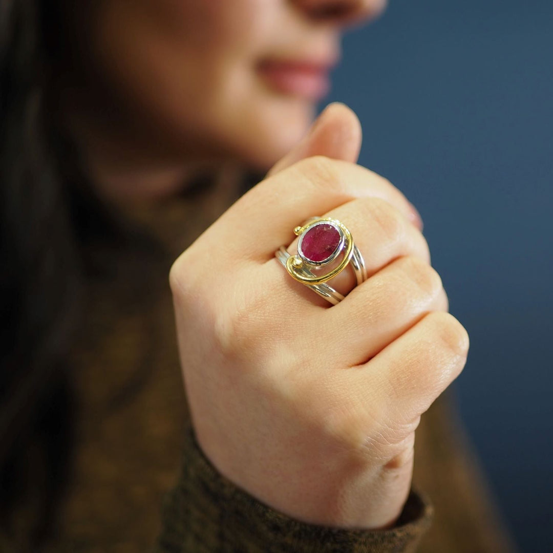 Close-up of a person holding their hand up, focusing on a Jasmine Ring in various gemstones by Gallardo & Blaine Designs adorned with a deep red gemstone. The background is blurred, highlighting the fine details of the jewelry.