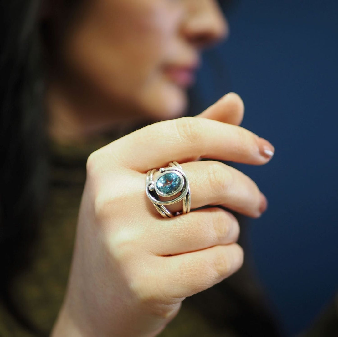 A close-up of a woman's hand displaying a Gallardo & Blaine Designs Jasmine Ring in various gemstones. Her fingernails are painted in a natural shade, and her face is blurred in the background. She wears a brown patterned top against a dark blue backdrop.