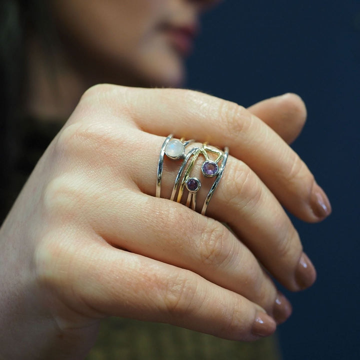 Close-up of a hand adorned with multiple Lattice Rings in various gemstones by Gallardo & Blaine Designs, featuring purple and white stones. The background is blurred, subtly showcasing the person's face and dark hair. The nails are neatly manicured with a natural polish, highlighting the contemporary design of each piece.