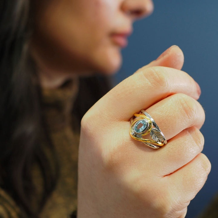 A close-up of a person holding up their hand, displaying an elegant Leaf Ring in Silver Gold & various gemstones by Gallardo & Blaine Designs with a blue stone set in an intricate design. The background is blurred, with the person's face out of focus.