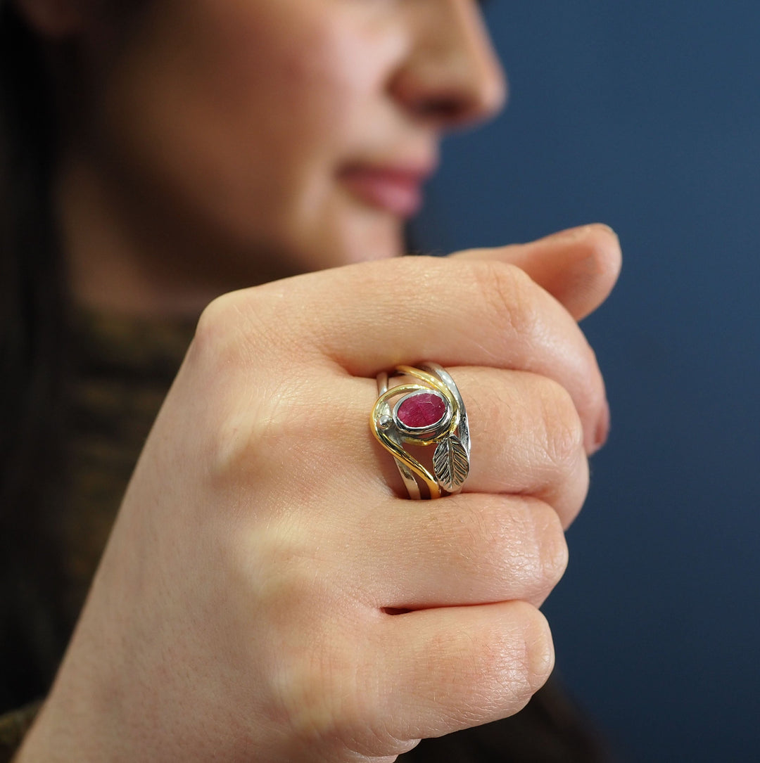 A close-up of a person's hand displaying the Gallardo & Blaine Designs Leaf Ring in Silver Gold & various gemstones, with a red gem and intricate leaf design in silver and gold. The person is partially visible in the background with a blurred face and dark hair. The primarily blue backdrop enhances the ring's beauty.