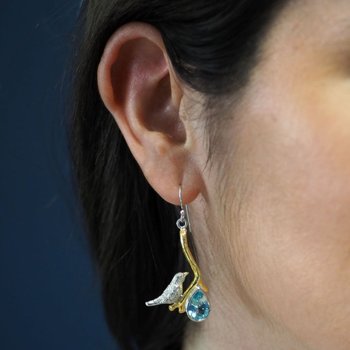 Close-up of a woman's ear wearing unique statement earrings. The earring features a small bird figure perched on a curved, gold-toned branch with a blue teardrop gemstone hanging below. The background is a simple, solid color. She is wearing the Lovebird Earrings in various gemstones by Gallardo & Blaine Designs.