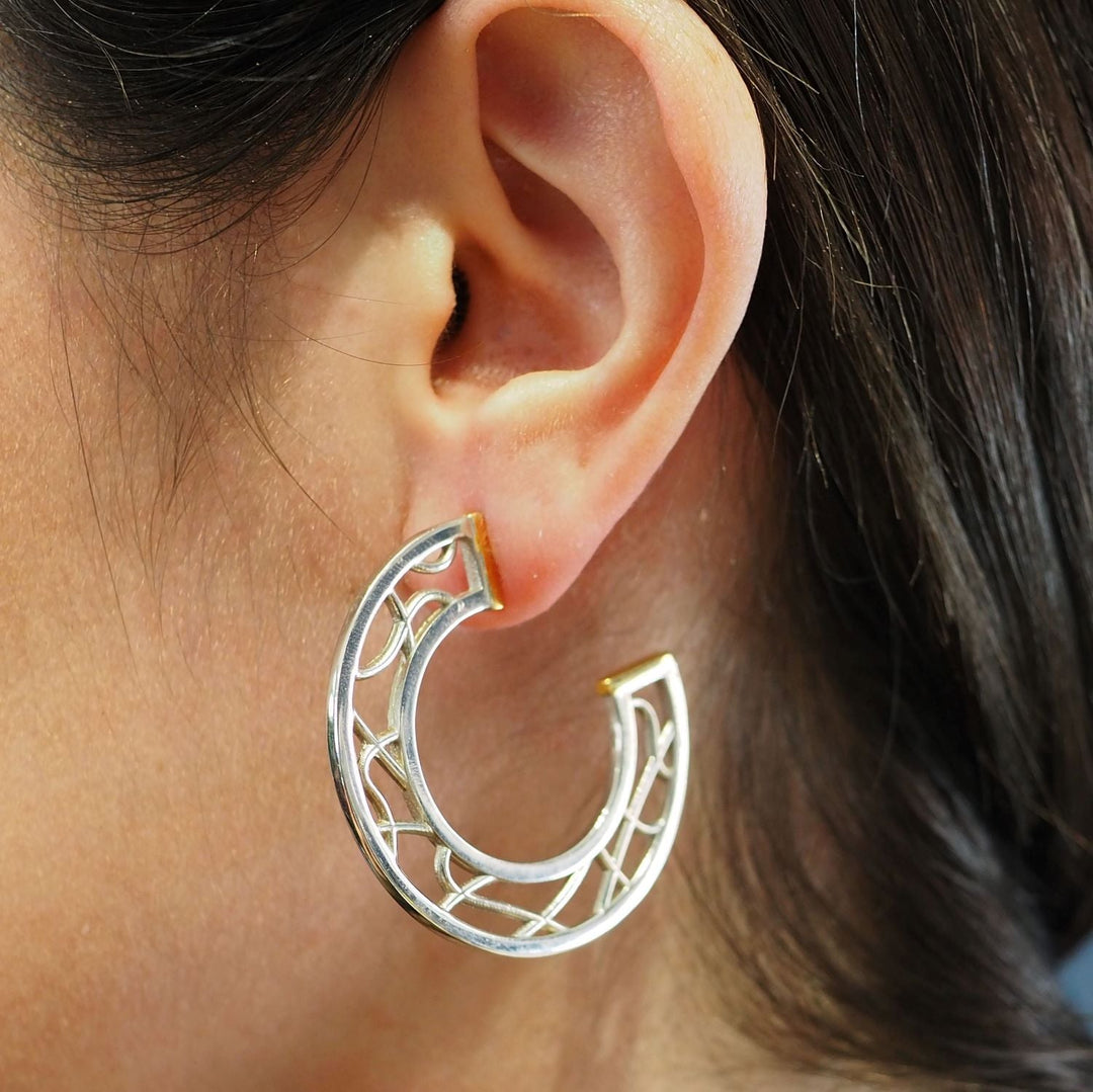 Close-up of a person's ear wearing a sterling silver hoop earring with an intricate, open lattice design. The Maya Earrings by Gallardo & Blaine Designs are shaped in a half-circle style and have a smooth, polished finish. The person's dark hair is partially visible.