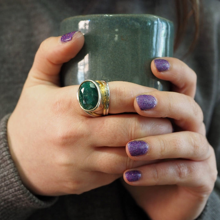 A person with glittery purple nail polish holds a green mug with both hands. They are wearing a large sterling silver & gold Party Ring in various gemstones by Gallardo & Blaine Designs on one hand and another adjustable ring on their middle and ring fingers. The person is dressed in a dark sweater.