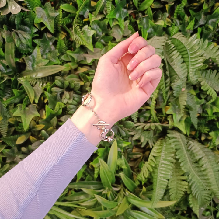 A hand wearing a Sea Honey Bracelet Silver by Gallardo & Blaine Designs featuring thick silver links and a clasp shaped like handcuffs is extended in front of a green leafy background. The arm is clothed in a long-sleeved, light purple top, showcasing the boldness of new designs.