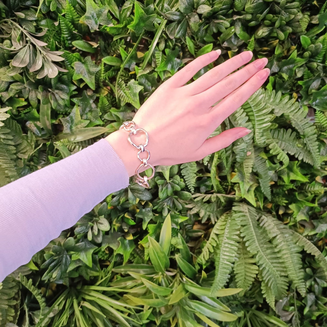 A person's hand, wearing a Sea Honey Bracelet Silver by Gallardo & Blaine Designs with thick silver links, is extended in front of a vibrant green leafy background. The sleeve of a light purple top is visible on the wrist.