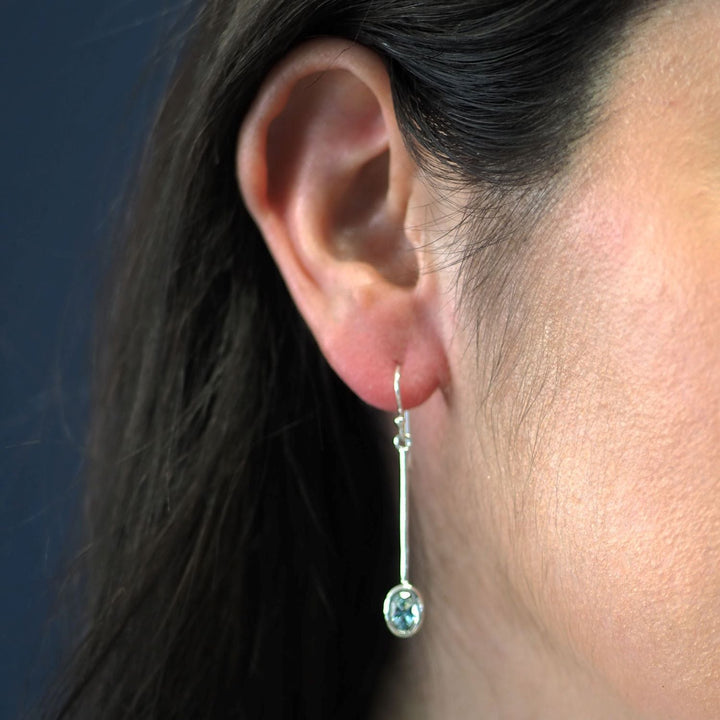 Close-up of a woman's ear adorned with Sequola Earrings in various gemstones by Gallardo & Blaine Designs, featuring a long sterling silver design that ends in a circular, light blue gemstone. The background is a blurred dark blue, highlighting the features of these exquisite gemstone dangle earrings and the woman's skin tone.