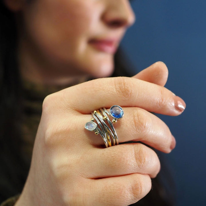 Close-up of a woman's hand wearing a stack of rings with blue gemstones, including a unique Gallardo & Blaine Designs Snake Ring in Silver Gold & various gemstones, held up to her face. The background is blurred, emphasizing the rings and the intricate detail of the stones. The woman has long dark hair and is seen against a blue backdrop.