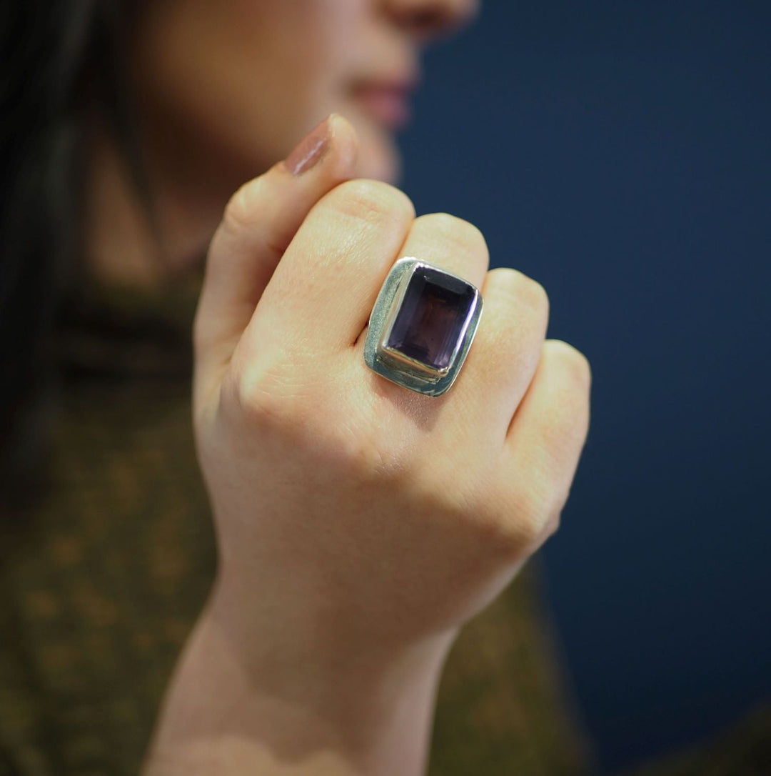A person with light brown nail polish displays their hand near their face, highlighting Gallardo & Blaine Designs’ Top Hat Ring in various gemstones against a blurred blue background.