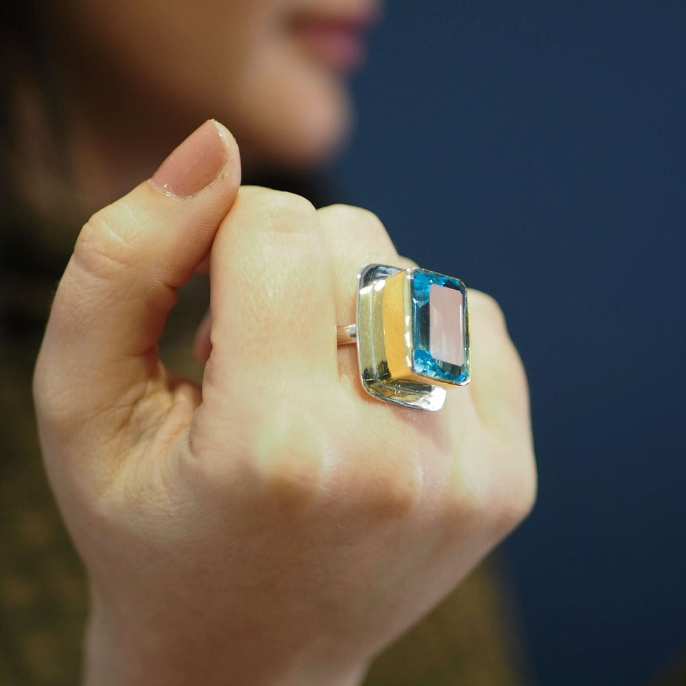 A close-up of a hand wearing the Gallardo & Blaine Designs Top Hat Ring, featuring a large rectangular blue gemstone set in sterling silver, with the wearer's face blurred in the background.