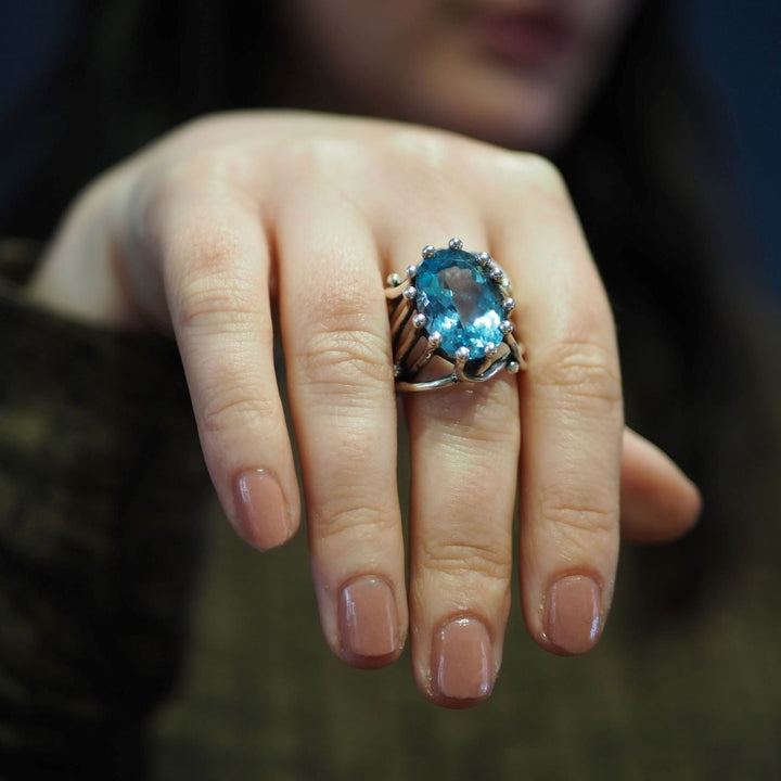 A close-up of a person's hand adorned with a chunky statement gemstone ring, the Willow Ring in various gemstones by Gallardo & Blaine Designs, featuring a prominent blue gemstone. The person's nails are neatly manicured with nude polish. The background is blurred, drawing attention to the adjustable ring.