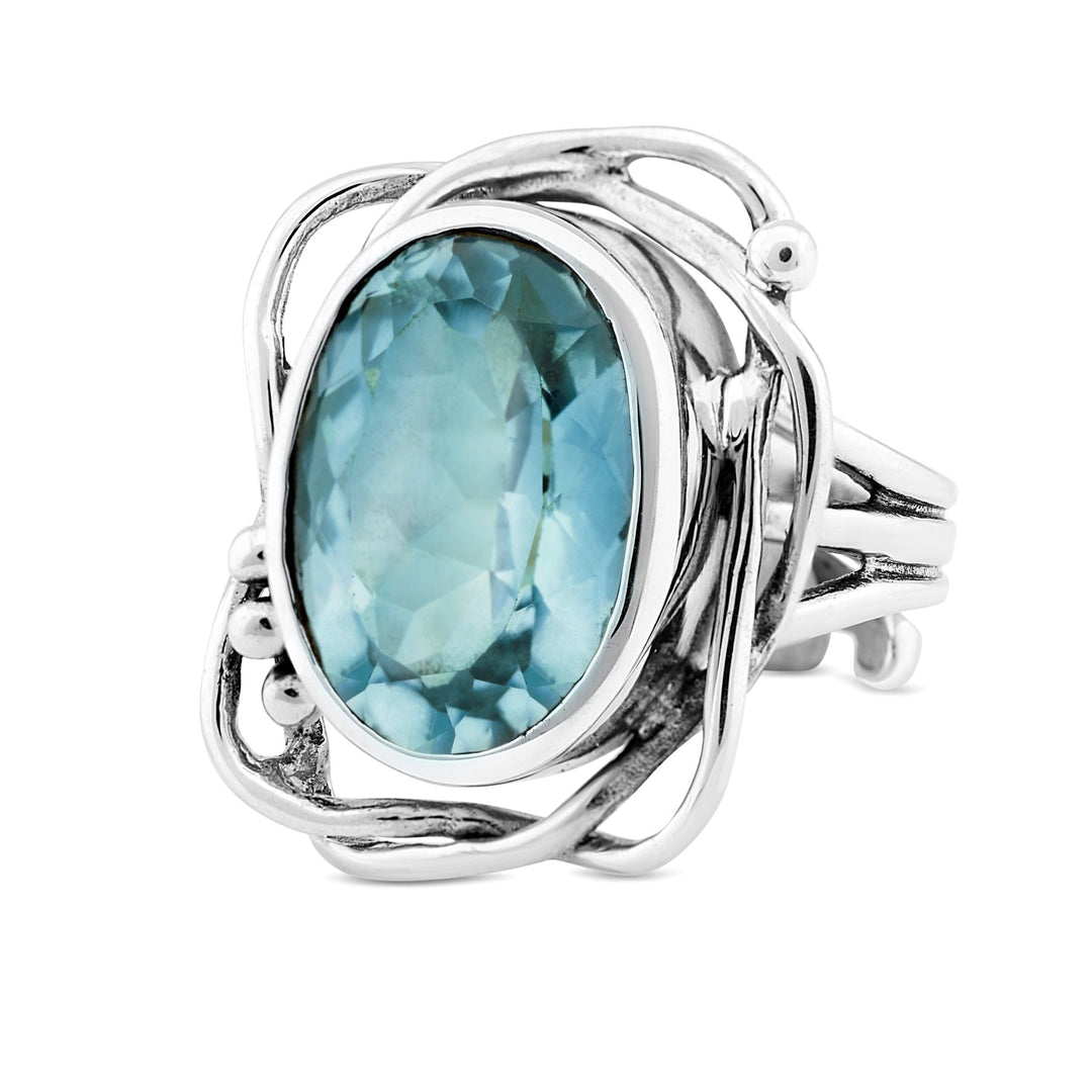 A silver ring with an abstract, wavy design features a large, oval-shaped blue gemstone at its center. The intricate silver setting wraps around the gemstone, giving this Gallardo & Blaine Designs Whirlpool Ring in various gemstones a modern and artistic appearance.