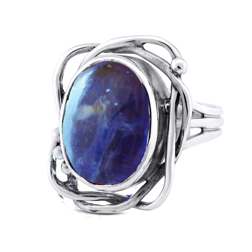 This Whirlpool Ring in various gemstones by Gallardo & Blaine Designs features an intricate design with intertwining bands and an oval-shaped blue gemstone in the center. The gemstone has varied shades of blue, giving it a unique appearance. The metalwork surrounding the gemstone adds a touch of elegance.