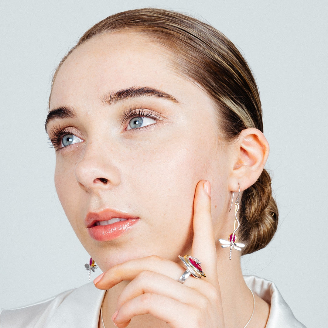 A contemplative person with slicked-back hair wearing a white shirt, Willow Earrings in Various Gemstones by Gallardo & Blaine Designs, and a matching ring with a gemstone. They are resting their chin on their fingers, gazing upward with a serene expression against a plain white background.