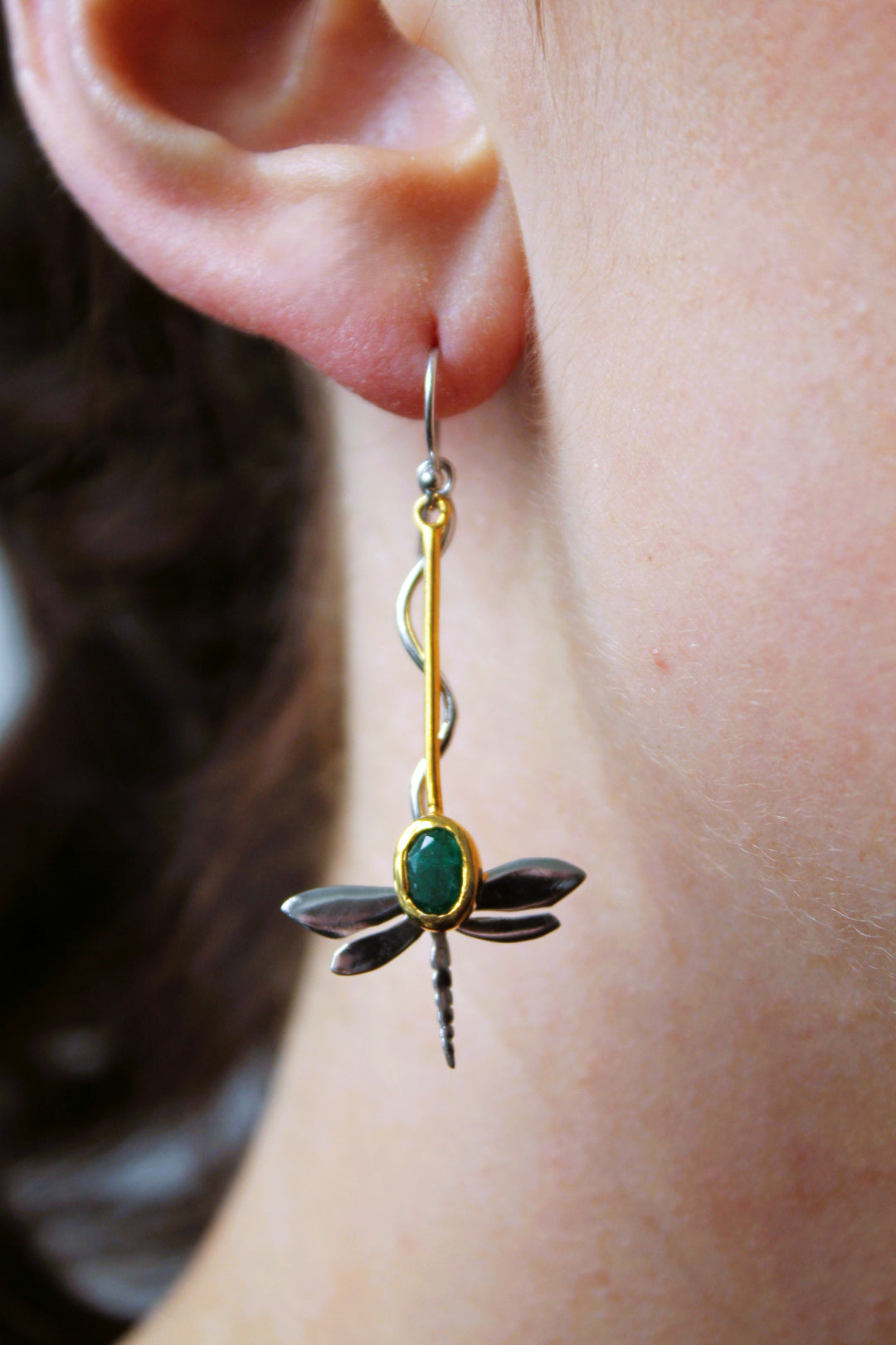 A close-up of a person’s ear wearing Gallardo & Blaine Designs’ Willow Earrings in Various Gemstones, featuring silver dragonfly dangles with gold and silver details and a green gem at the center.