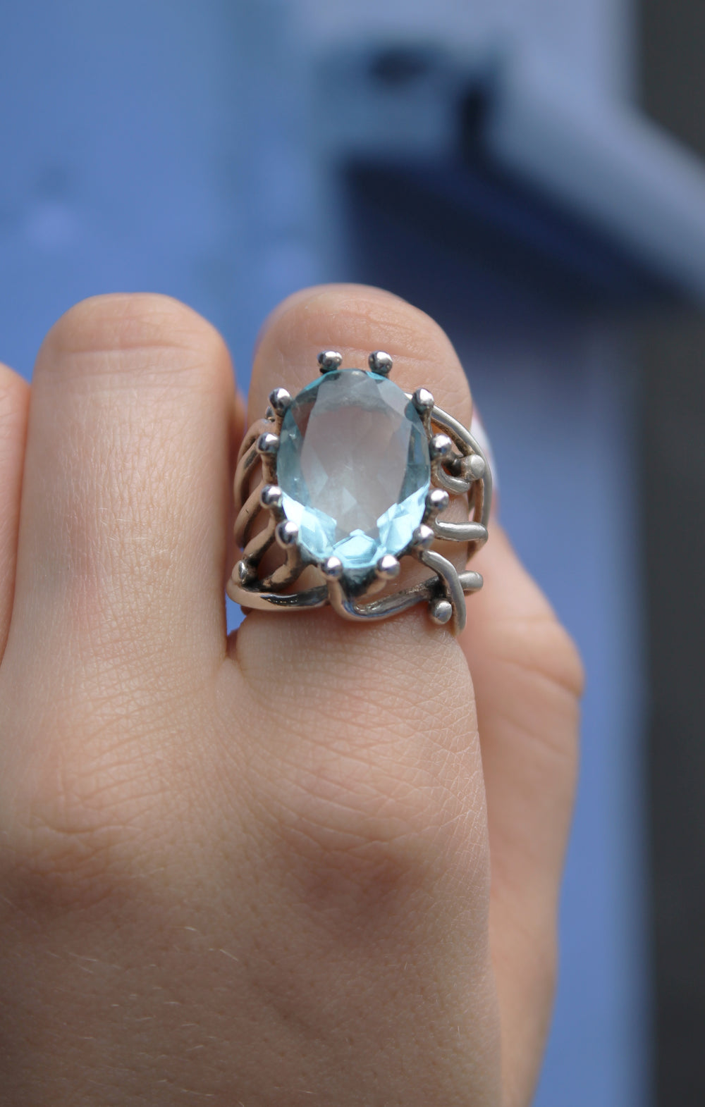 A close-up of a hand wearing the Willow Ring in Blue Topaz by Gallardo & Blaine Designs, a sterling silver piece with an ornate, organic design. The background features soft, soothing blue tones.
