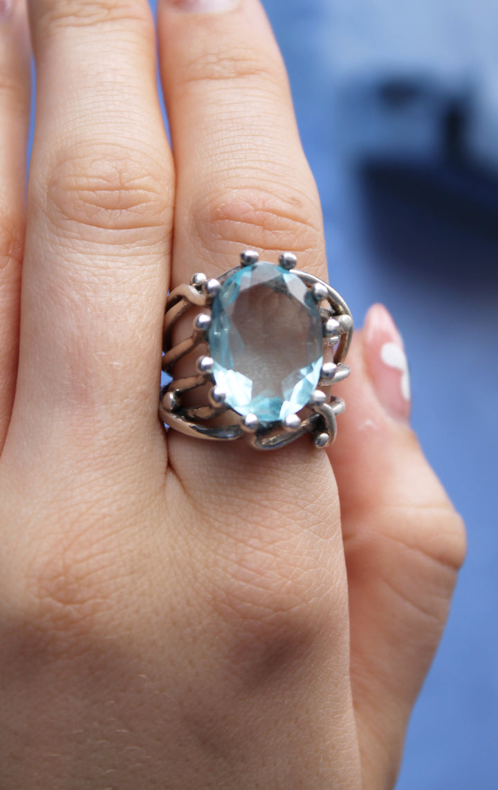 A close-up of a hand wearing the Gallardo & Blaine Designs Willow Ring, featuring a striking Blue Topaz set in an ornate sterling silver band, shown against a soft blue background.