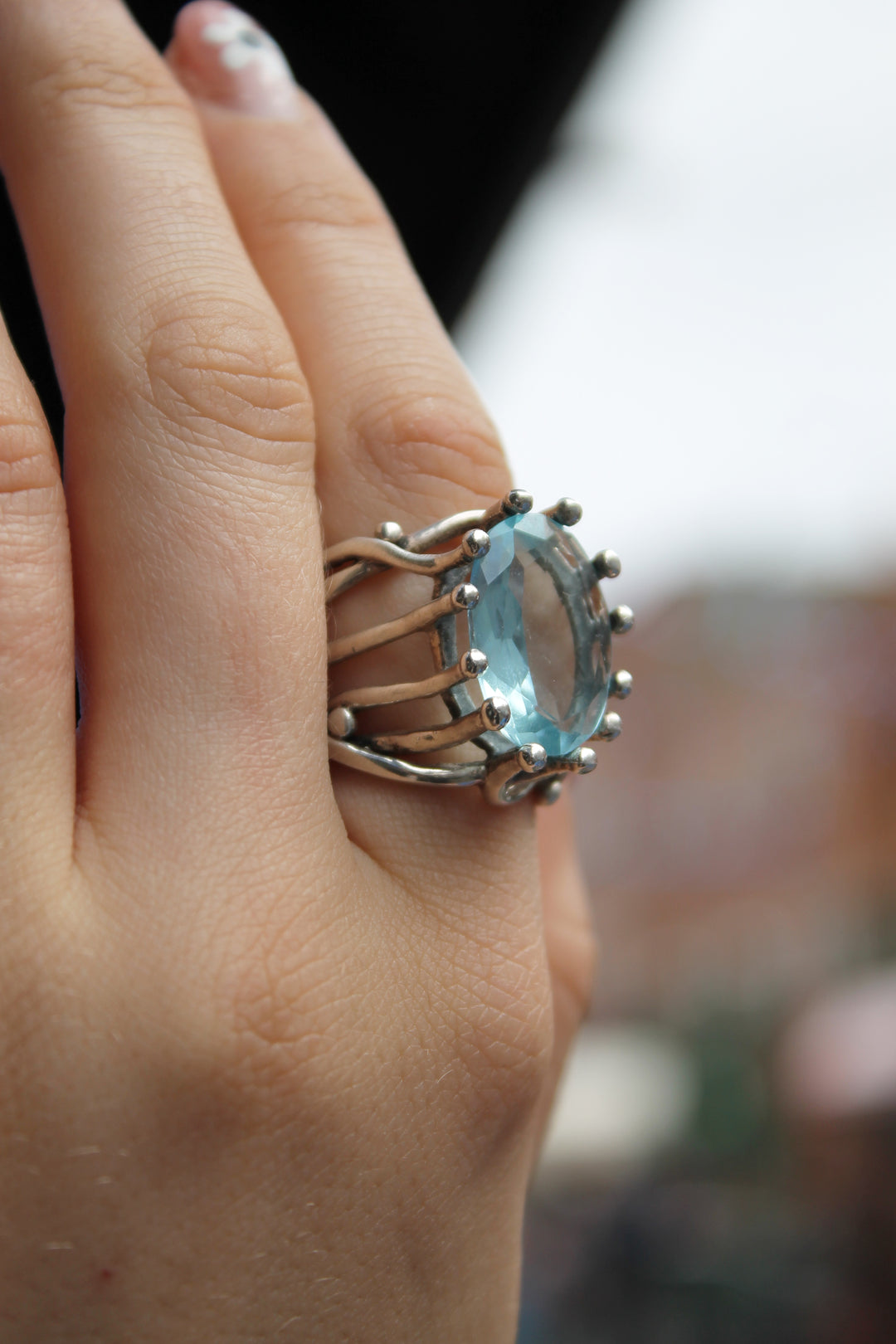 A hand showcases the Gallardo & Blaine Designs Willow Ring in sterling silver with an oval Blue Topaz gemstone, set in decorative prongs, against a blurred outdoor background.
