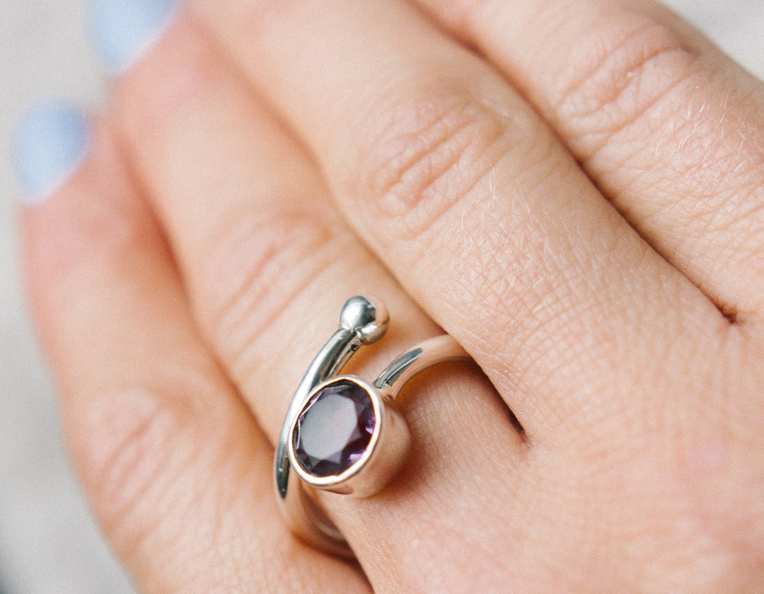 Close-up of a hand with light blue painted nails wearing the Honeysuckle Ring in Silver Gold & gemstones by Gallardo & Blaine Designs. The unique silver design features an open-ended band with a shiny round purple gemstone in a minimalist bezel setting on one end and a smooth round bead on the other.