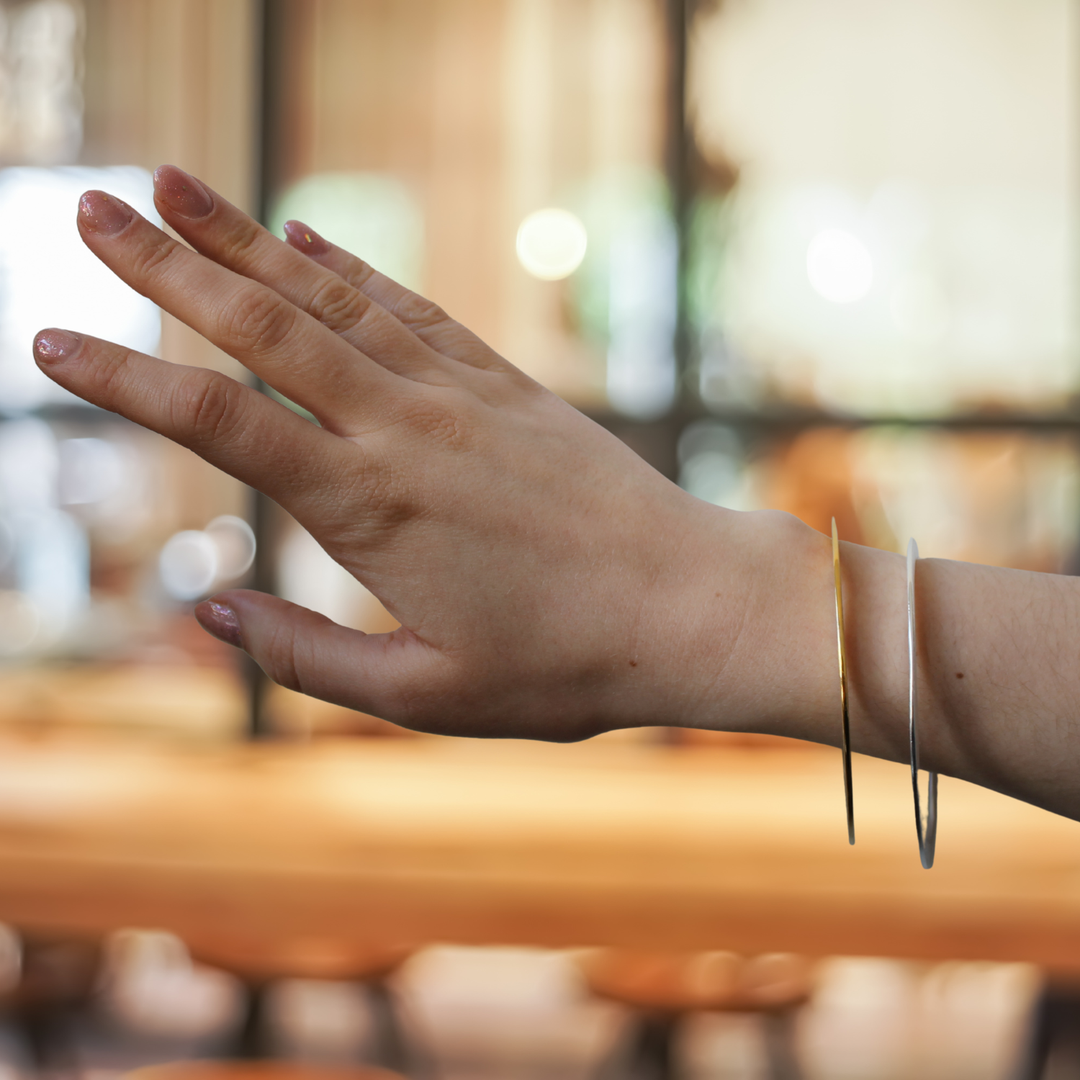 A hand with light pink nails wears Gallardo & Blaine Designs’ Flat Satin Bangles in sterling silver and gold vermeil, both thin and metallic, against a softly lit indoor backdrop.