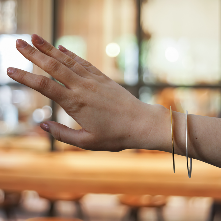 A hand with light pink nails wears Gallardo & Blaine Designs’ Flat Satin Bangles in sterling silver and gold vermeil, both thin and metallic, against a softly lit indoor backdrop.