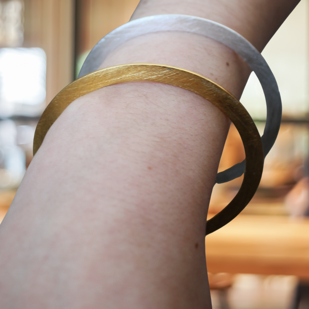 A close-up shows a person's arm wearing two overlapping Gallardo & Blaine Designs Flat Satin Bangles in silver and gold, set against a softly blurred indoor background with warm lighting.