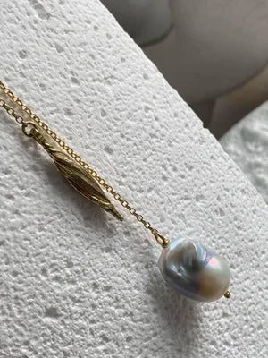 The Gathered Forms Lariat Necklace by Inner Island, crafted in gold vermeil with a leaf-shaped charm and Baroque pearl pendant, rests elegantly on a textured white surface.