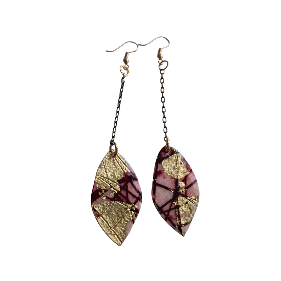 Introducing the Rothlú Gile Abstract Leaf Batik Textile Earrings in Red/Gold: a stunning pair of dangling earrings featuring leaf-shaped pendants with a unique texture and an abstract gold and maroon design. Each earring is connected to the hypo-allergenic gold-plated hook via a black chain link, elegantly contrasting against the white background.